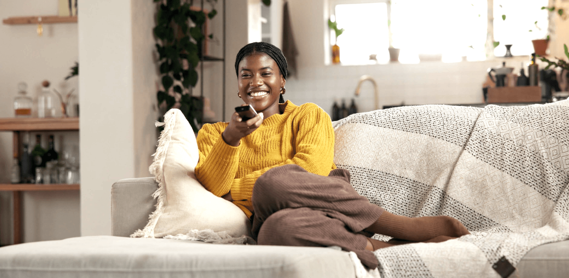 A smiling woman in a yellow sweater relaxes on a couch, holding a TV remote and looking toward the camera.