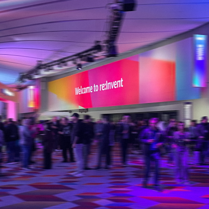 A photograph of a large, crowded convention center hall. A large, curved sign with a colorful pink, orange, and purple gradient background reads "Welcome to re:Invent". The space is illuminated with purple and blue lights, and the floor has a geometric pattern. Numerous attendees are walking around the hall.