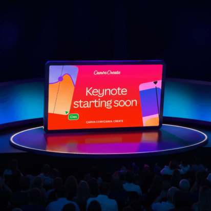 A wide shot of a tech conference stage featuring a large, tablet-shaped central screen that reads "Canva Create" and "Keynote starting soon." The screen is bright red, flanked by curved, glowing blue side panels. In the foreground, a large audience is seated in a darkened theater setting, facing the stage.