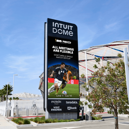 A tall digital billboard outside the Intuit Dome displaying a FIFA World Cup advertisement featuring soccer player Lionel Messi in action. The screen reads "ALL MEETINGS ARE FLEXIBLE" above the dates June 11 through July 19, alongside logos for Fox and various corporate sponsors like Patron and US Bank. The sign stands on a sunny, palm-tree-lined sidewalk with the stadium's exterior visible in the background behind a white fence.