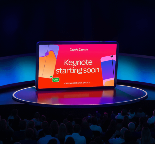 A wide shot of a tech conference stage featuring a large, tablet-shaped central screen that reads "Canva Create" and "Keynote starting soon." The screen is bright red, flanked by curved, glowing blue side panels. In the foreground, a large audience is seated in a darkened theater setting, facing the stage.