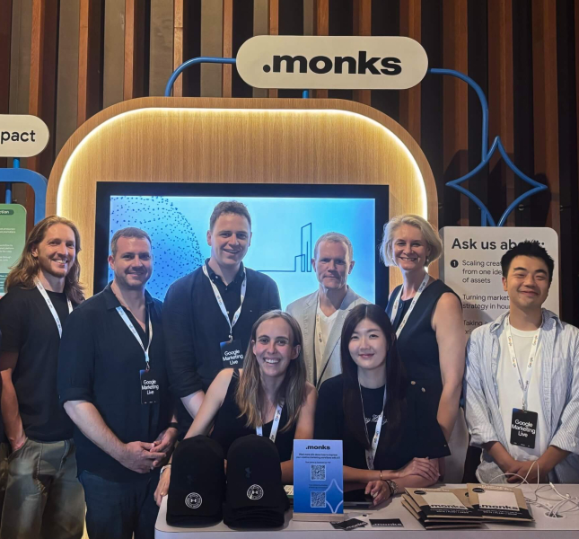 A group of ten smiling professionals posing together at a ".monks" event booth. The attendees are wearing lanyards with "Google Marketing Live" badges. Behind them is a large display screen, an illuminated ".monks" logo, and booth signs reading "Brand impact" and "Ask us about:". In the foreground, a table is set up with branded black caps, brochures, and a small sign featuring a QR code.