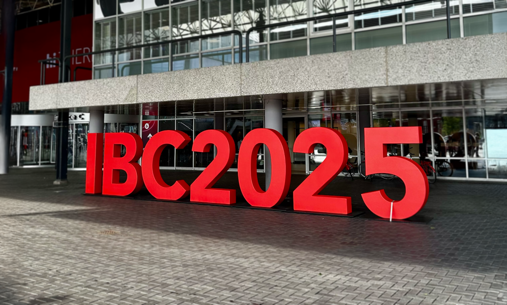 Large, three-dimensional red letters spelling "IBC2025" stand on a brick plaza in front of the entrance to a modern convention center with a glass facade.
