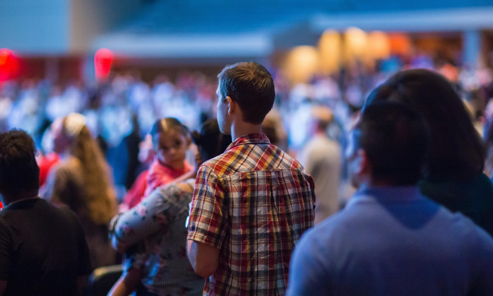 A rear view of a standing crowd in a large, dimly lit indoor venue, focusing on a man in a plaid shirt in the center. To his left, a woman holds a small child who is looking sideways, while the foreground and background contain other attendees blurred out of focus.