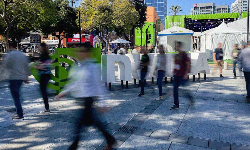 A wide-angle, slightly blurred shot of an outdoor plaza at the NVIDIA GTC 2026 conference in San Jose. Large, 3D white letters spelling out "NVIDIA" stand in the center, with the green NVIDIA logo to the left. People are captured in motion, appearing as blurred figures walking across the stone-tiled ground, creating a sense of a busy, electric atmosphere. In the background, there are green banners, white event tents, trees, and city buildings under a clear blue sky.